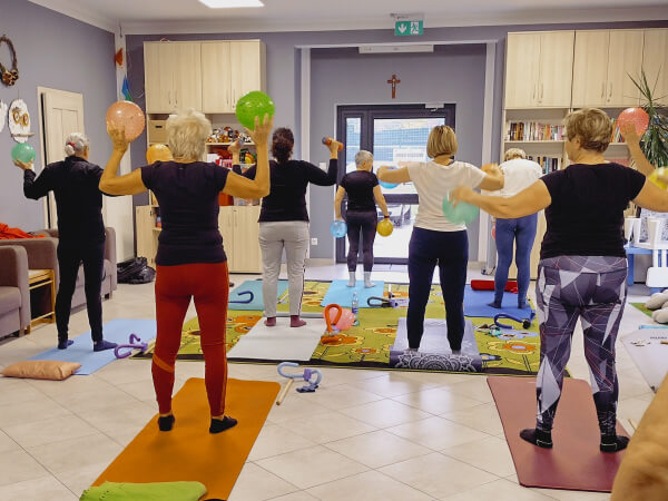 Zajęcia z gimnastyki. Pełna sala dorosłych, którzy stoją na matach i trzymają w dłoniach piłeczki.