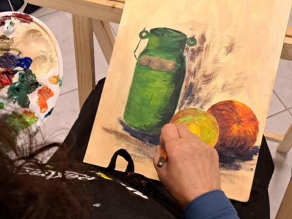 Dziewczyna maluje obraz na którym jest zielona doniczka, mała dynia i cytryna. Maluje farbami akrylowymi.