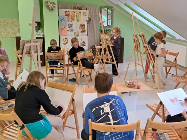 Dzieci stoją przed sztalugami lub siedzą na krzesłach w kręgu malując martwą naturę.