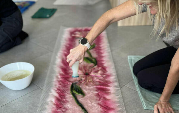 Uczestniczka jest w takcie tworzenia szala szesankowego. Szal jest różowy z motywem kwiatowym.